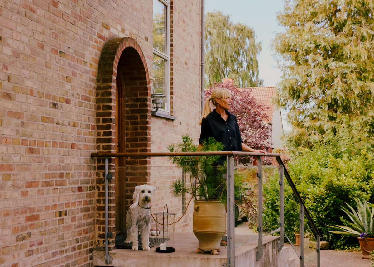 Kvinde og hund på trappe foran gulstenshus med have og hæk